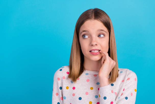 Photo of adorable unsure small girl wear dotted shirt biting finger looking empty space isolated blue color background. - Фото, зображення