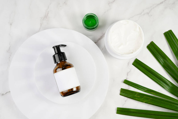 A mock-up of a brown cosmetics bottle with dispenser and white label on white round podium, round cream container on marble background and green palm leaf, top view - Foto, imagen