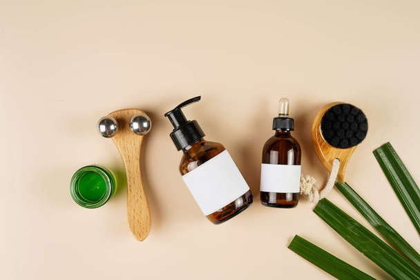 A mock-up of a brown cosmetics bottle with dispenser and white label, round green cream container, bottle with pipette, face brush, face massage roller on beige colored background lined up, top view - Photo, Image