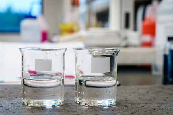 Два стакана с прозрачными жидкостями и чистыми этикетками на блокноте в лаборатории - Фото, изображение