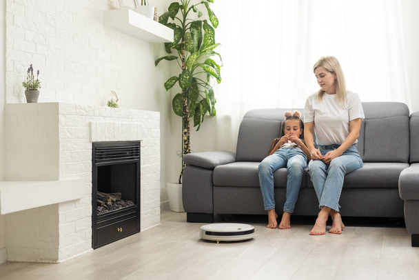 Cute child near mother looking at robotic vacuum cleaner in living room. - Zdjęcie, obraz
