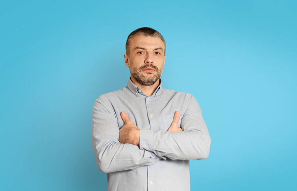 Studio Shot Of Serious Middle Aged Male Posing Crossing Hands Looking At Camera Standing On Blue Background. Self Confidence And Entrepreneurship Concept - Foto, imagen