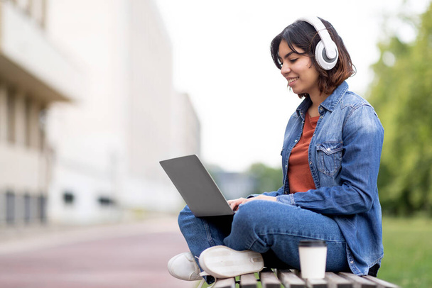 Smiling arab female student in wireless headphones study with laptop outdoors, young middle eastern woman sitting with computer on bench at campus, enjoying distance learning, copy space - Фото, изображение