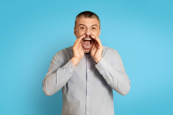 Emotional Middle Aged Male Shouting Announcing Offer Holding Hands Near Mouth Looking At Camera Standing Posing Over Blue Background. Studio Shot Of Man Yelling Aggressively - Foto, immagini