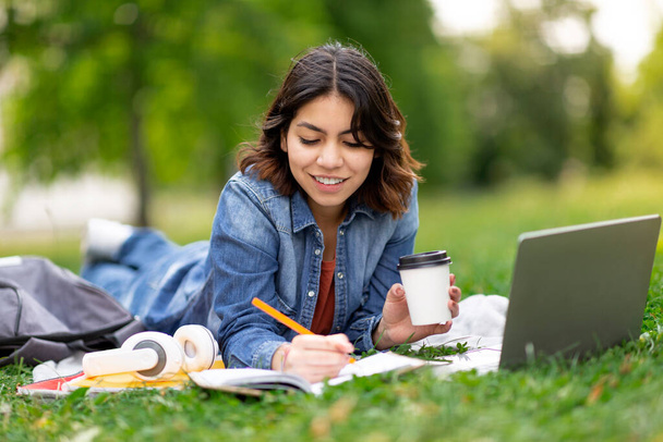 Молодая арабская студентка с ноутбуком и блокнотом в парке, улыбающаяся ближневосточная женщина лежит на лужайке, пьет кофе и пишет в блокноте, готовится к экзамену на улице - Фото, изображение