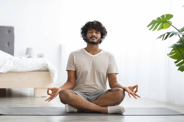 Мирный индийский мужчина тысячелетия в домашней одежде практикующий йогу дома, сидящий на коврике с закрытыми глазами, медитирующий, копирующий пространство, наслаждающийся утренней тренировкой, полноразмерный снимок. Концепция снятия напряжения - Фото, изображение
