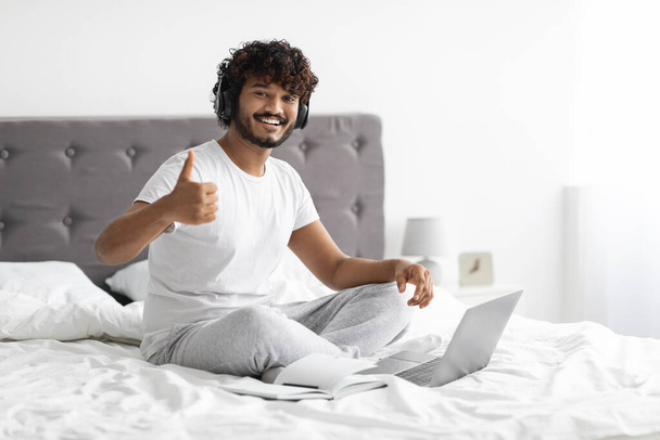 Positive curly millennial eastern guy sitting on bed, using wireless headset and laptop, showing thumb up and smiling, attending webinar on Internet, bedroom interior, copy space - 写真・画像