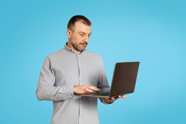 Средневековый бизнесмен с помощью портативного компьютера, работающего онлайн позируя в студии на синем фоне. Концепция внештатной карьеры и интернет-технологий - Фото, изображение
