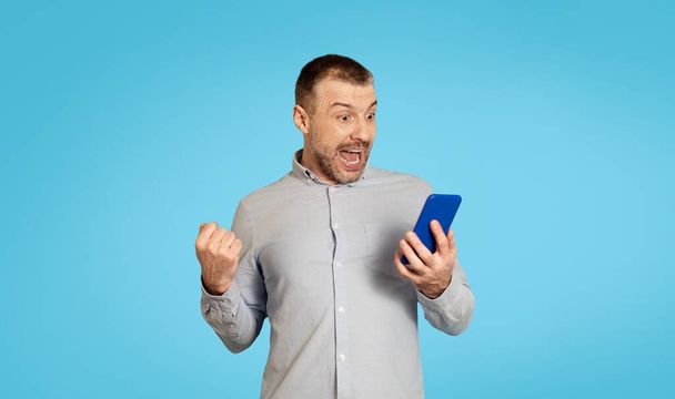 Emotional Middle Aged Man Holding Phone Shaking Fists Standing Over Blue Studio Background. Male With Cellphone Gesturing Yes Celebrating Success. Great Mobile Application And Offer - Foto, Bild