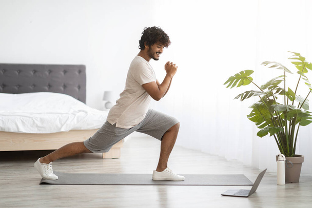 Athletic young indian guy in sportswear exercising at home, working on legs strength, looking at laptop screen and doing workout, enjoying online fitness video, copy space, bedroom interior, panorama - Fotó, kép