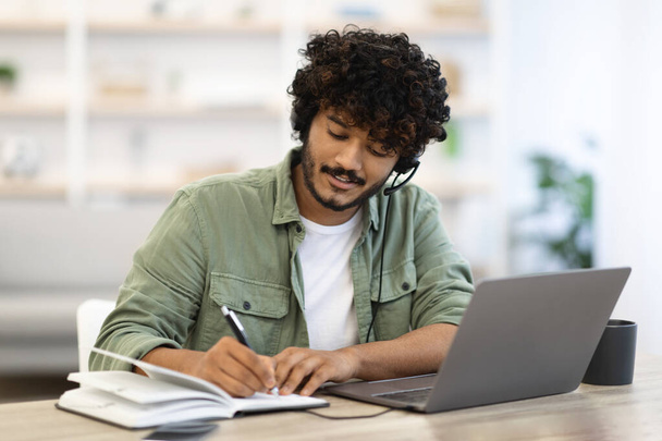 Young indian man having online training or attending webinar, sitting at workdesk, using laptop and headset, taking notes and smiling, home interior, copy space. Online education concept - Photo, Image