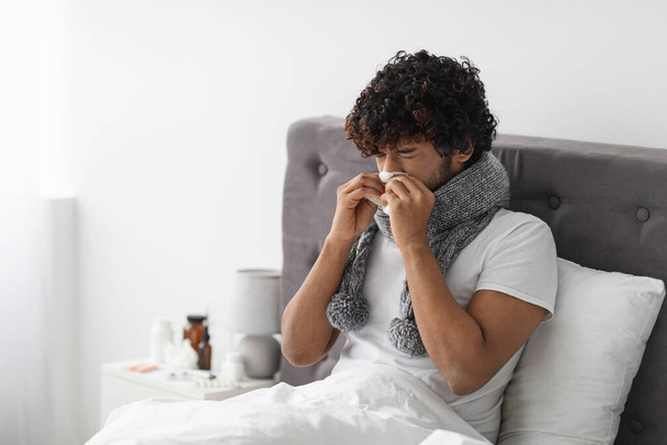 Sick millennial eastern guy with scarf around his neck staying in bed at home, using tissue, sneezing nose, curly young man suffering from coronavirus, copy space. Flu, COVID-19 concept - Foto, Imagem