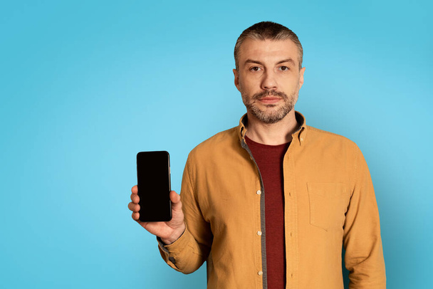 Серьезный мужчина средних лет показывает смартфон с пустым экраном, позирующий в студии на синем фоне. Мужское рекламное мобильное предложение. Коммуникации, технологии и гаджеты. Mockup - Фото, изображение