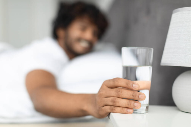 Selective focus on glass of fresh spring water next to eastern guy bed, unrecognizable indian man smiling and taking glass of water after waking up in the morning, copy space. Hydration concept - Photo, Image