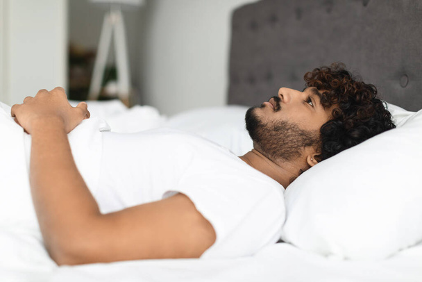 Closeup of depressed young eastern guy lying in bed alone, staring at the celining, young hindu man feeling down, side view, copy space. Mental health problems in millennials concept - Foto, afbeelding