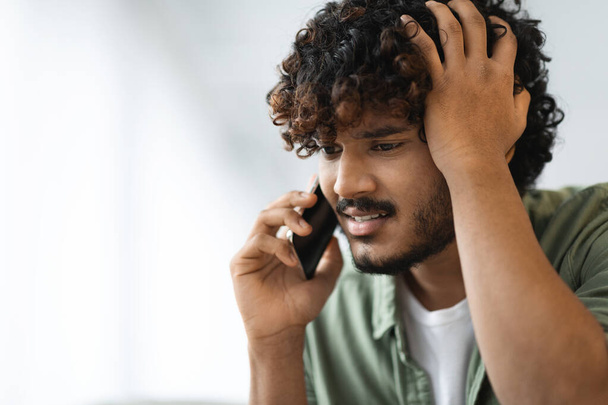Worried young curly eastern man having phone conversation, unhappy indian guy talking on smartphone and touching his head, having bad news, blurred background, closeup, copy space - Foto, imagen