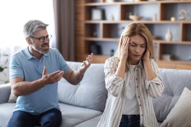 Sad upset angry adult european husband yells at unhappy wife, covers ears in living room interior. Relationship problems, quarrel, stress, crisis, scandal, family conflict and human emotions at home - Photo, Image