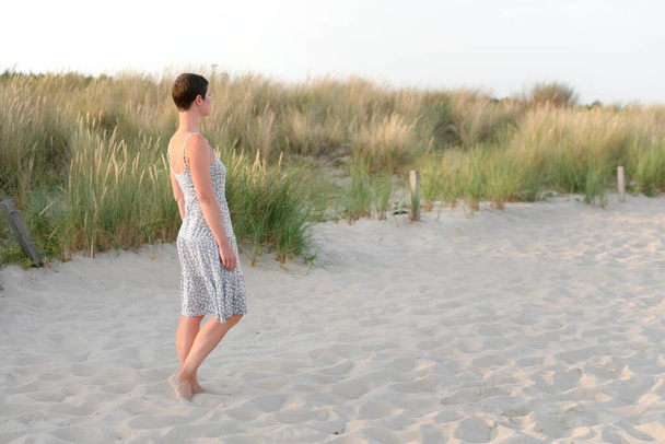 Attractive short haired woman in her thirties, dressed in light summer dress standing barefoot on the beach with grass in the background. Concept for vacation, relaxation, mental health - Fotografie, Obrázek