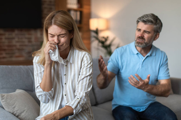 Angry upset mature european husband yells at crying wife, couple quarreling in living room interior. Human emotions, relationship problems at home, stress, pressure, scandal and domestic violence - Photo, Image