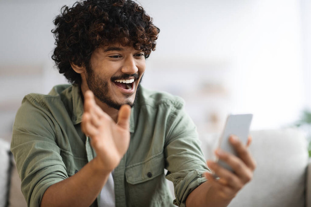 Emotional young hindu guy having online party with friends while staying home, sitting on couch in living room, using smartphone, waving and smiling at cell phone screen, closeup, copy space - Foto, Imagen