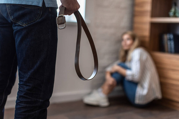 Angry aggressive european husband with belt threatens frightened wife with belt, lady sits on floor in living room interior, cropped. Conflict, pressure, domestic violence, abuse in marriage at home - Photo, image