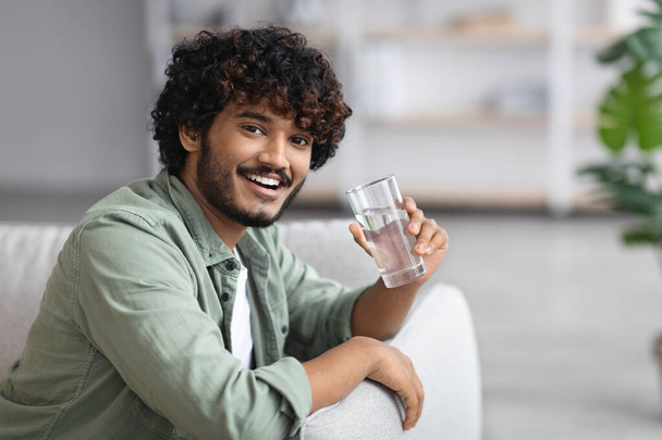 Handsome dark-skinned young man drinking water and smiling at camera, cheerful indian guy sitting on couch in living room, holding glass of fresh water, chilling alone at home, copy space - Photo, image