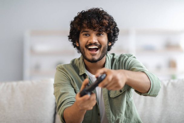 Young happy indian man laughing and playing video games on weekend, sitting on couch alone at home, holding joystick, having fun, copy space. Modern technologies and entertainment concept - Foto, afbeelding