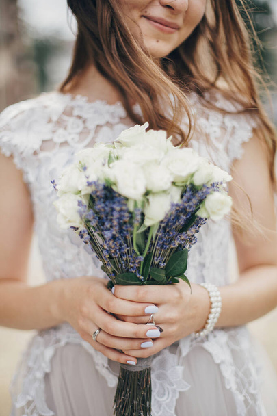 Portrait of sensual bride with wedding bouquet of roses and lavender in stylish gown. Provence wedding. Beautiful bride in stylish vintage dress posing in sunny street of european city - Foto, Imagen