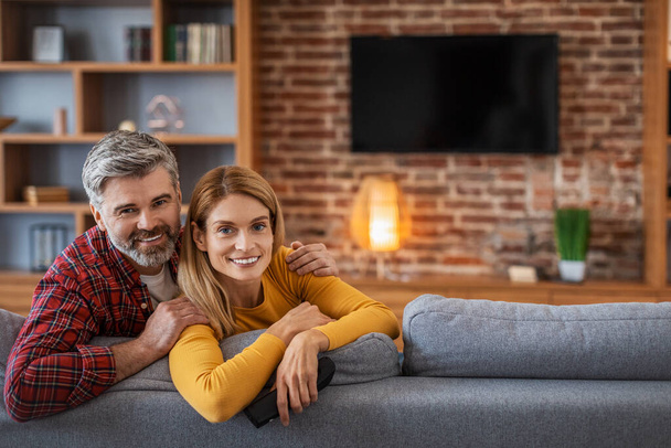 Happy adult european man and woman looking at camera, enjoy relaxing in living room interior with tv with blank screen. Free time in cozy home, love and relationships, news, movies in evening together - Foto, Bild