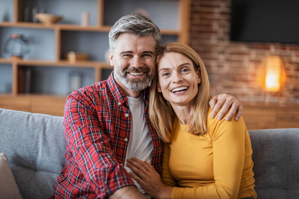 Smiling middle aged handsome european male and female hugging and enjoy tender moment, look at camera, sitting on sofa in living room interior. Love, family relationships at home, people emotions - Фото, зображення