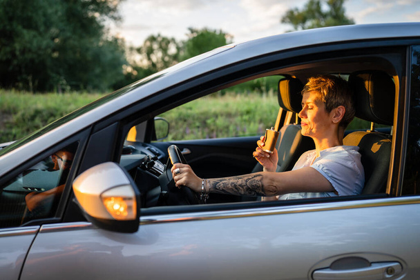 One woman mature caucasian female with tattoo on arms hold flask while sitting in the car alone driver on parked automobile drinking alcohol abuse and alcoholism concept copy space side view - Zdjęcie, obraz