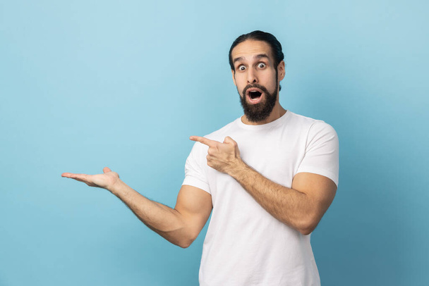 Portrait of astonished man with beard in white T-shirt presenting copy space on palm for commercial text, blank wall with idea presentation. Indoor studio shot isolated on blue background. - Photo, Image