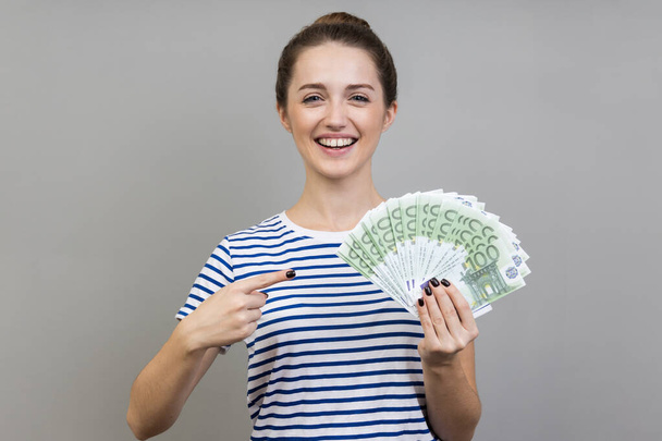 Portrait of woman wearing striped T-shirt pointing finger at fan of euros in her hand looking at camera with toothy smile, cash, money exchange service. Indoor studio shot isolated on gray background. - Photo, image