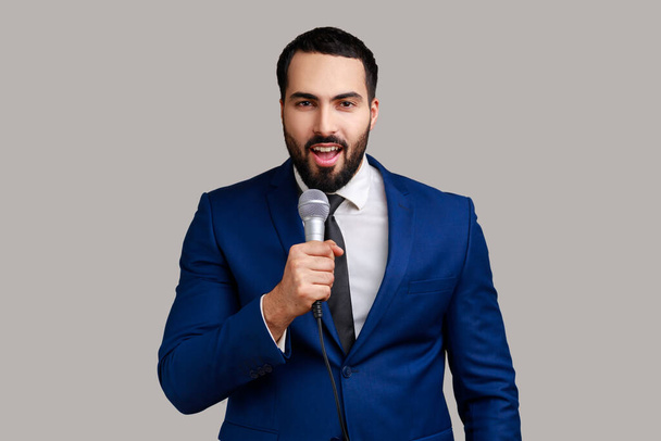 Positive handsome bearded man standing with microphone in hands, sing songs with happy optimistic expression, wearing official style suit. Indoor studio shot isolated on gray background. - Фото, зображення