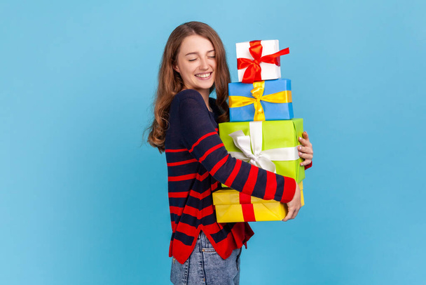 Attractive woman wearing striped casual style sweater, keeping eyes closed while embracing many present boxes, extremely happy to celebrate birthday. Indoor studio shot isolated on blue background. - Foto, immagini