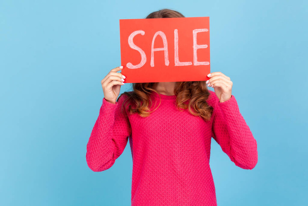 Portrait of unknown woman wearing pink pullover, covering face with Sale inscription, market discounts, black friday, low prices. Indoor her studio shot isolated on blue background. - Fotó, kép
