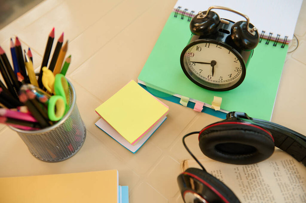 Still life. View from above with focus on a black alarm clock on open notepad with school supplies, audio headset and stationery on blurred foreground. Back to school. Education. Teachers day concept - Foto, afbeelding