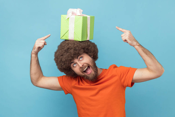 Portrait of positive funny man with Afro hairstyle wearing orange T-shirt holding gift box on his head and pointing with fingers, expressing excitement. Indoor studio shot isolated on blue background. - Foto, imagen