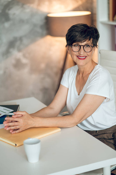 In the office. Dark-haired woman in eyeglasses sitting at the table in the office - Fotografie, Obrázek