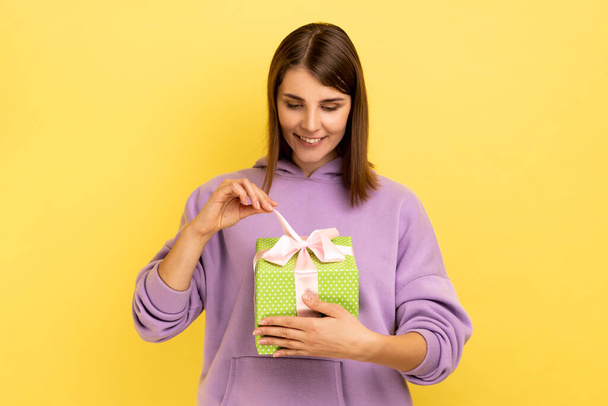 Young beautiful smiling woman feeling happy, opens present box with happy satisfied expression, being glad to get gift, wearing purple hoodie. Indoor studio shot isolated on yellow background. - Zdjęcie, obraz