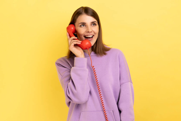 Portrait of beautiful young adult woman with excited expression talking on red retro telephone, holding handset, wearing purple hoodie. Indoor studio shot isolated on yellow background. - Photo, Image