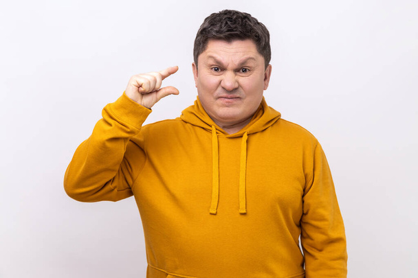 Portrait of funny man making little bit gesture with fingers and smiling sarcastic, showing minimum size, begging few more, wearing urban style hoodie. Indoor studio shot isolated on white background. - Foto, afbeelding