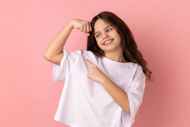 Portrait of smiling little girl wearing white T-shirt showing her arm muscle and point it with finger, proud of her strength and leadership skills. Indoor studio shot isolated on pink background. - Photo, image