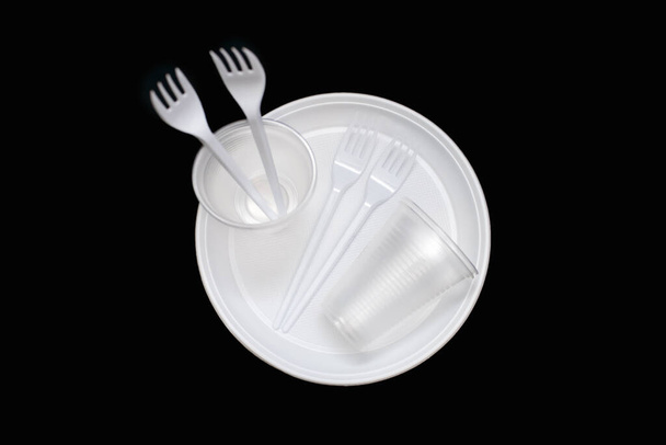 Одноразовая белая пластиковая посуда в виде тарелок, стаканов и вилок на черном фоне. Пластиковая посуда для пикника или вечеринки на открытом воздухе. Пластиковые загрязнения и экологические проблемы - Фото, изображение