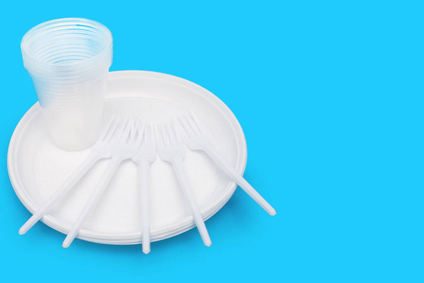 Одноразовий білий пластиковий посуд у вигляді тарілок, окулярів та виделки на синьому фоні. Вільний простір для тексту. Пластикове начиння для пікніка або вечірки на відкритому повітрі
 - Фото, зображення