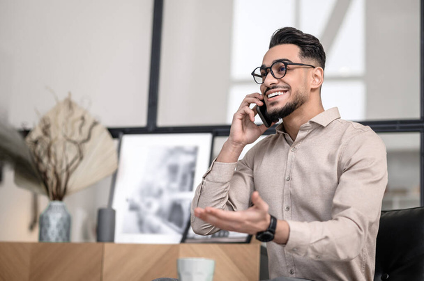 Phone call. Smiling young bearded elegant man gesturing talking on smartphone while sitting indoors - Φωτογραφία, εικόνα