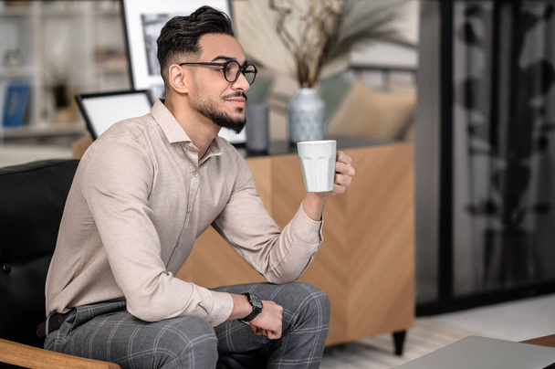 Morning coffee. Meditating young bearded stylish man with coffee sitting sideways to camera in room at home - Фото, зображення