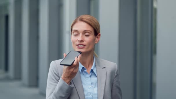 Outdoor portrait of confident successful business lady recording audio message for her assistant, talking to smartphone, standing near office building, slow motion, free space - Imágenes, Vídeo