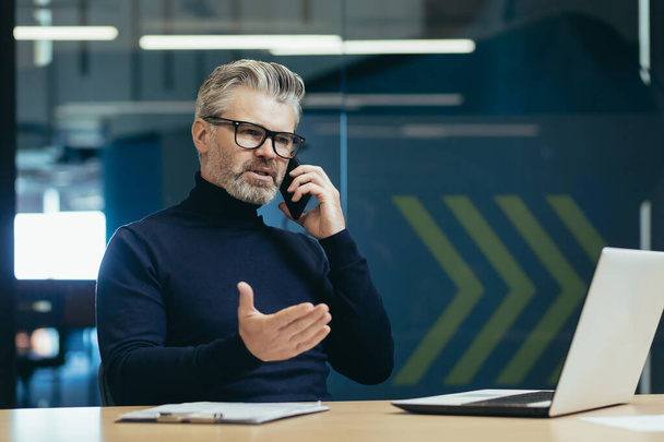 Senior and mature boss businessman working inside office building, man thoughtful and serious talking on the phone, gray haired investor in glasses working using laptop - Foto, immagini