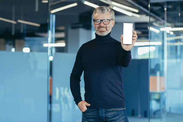 Senior experienced gray haired businessman working inside office, portrait of man with phone, mature investor smiling and looking at camera, showing white mobile phone screen - Foto, afbeelding
